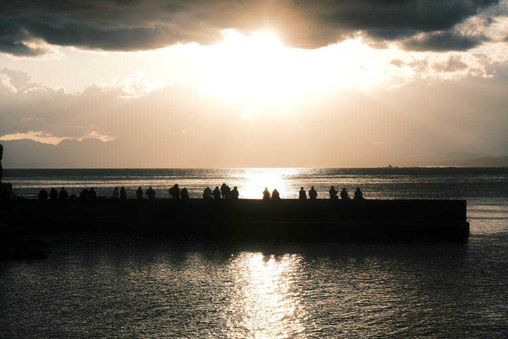 夕日に照らされた海と、堤防に並ぶ人たちのシルエット